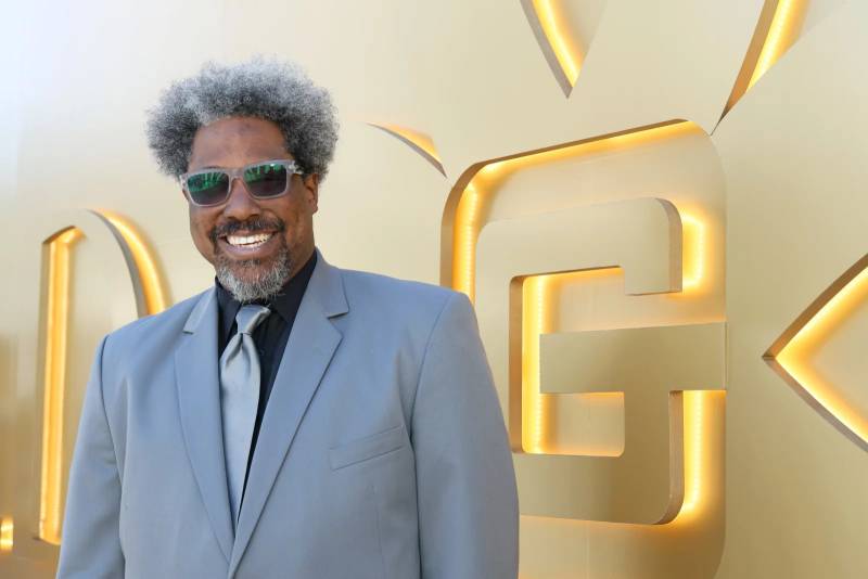 A middle-aged Black man in a light bluish greyish suit and sunglasses smiles against a yellow and gold backdrop