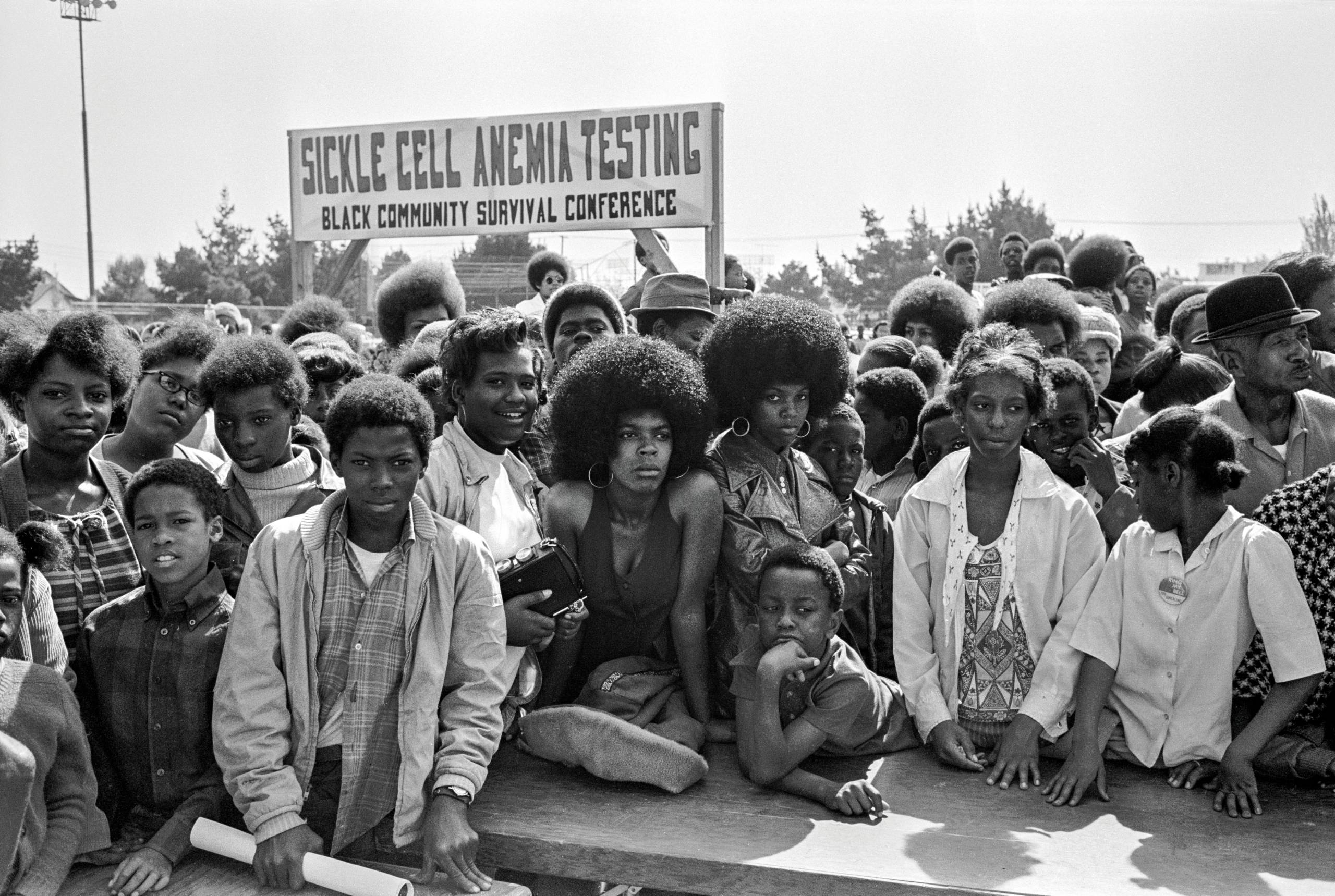 A black and white archival photo of a large group of African-American people gathered outside, behind them a sign reads "sickle cell anemia testing."
