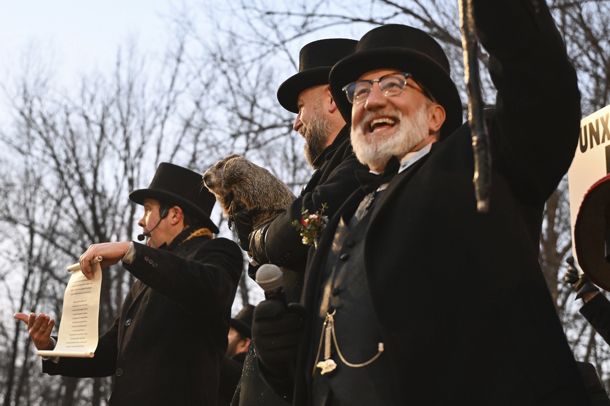 Three men in top hats and black winter coats stand in a line, outdoors, near a line of trees that are bare for the winter. The middle man is holding a groundhog.