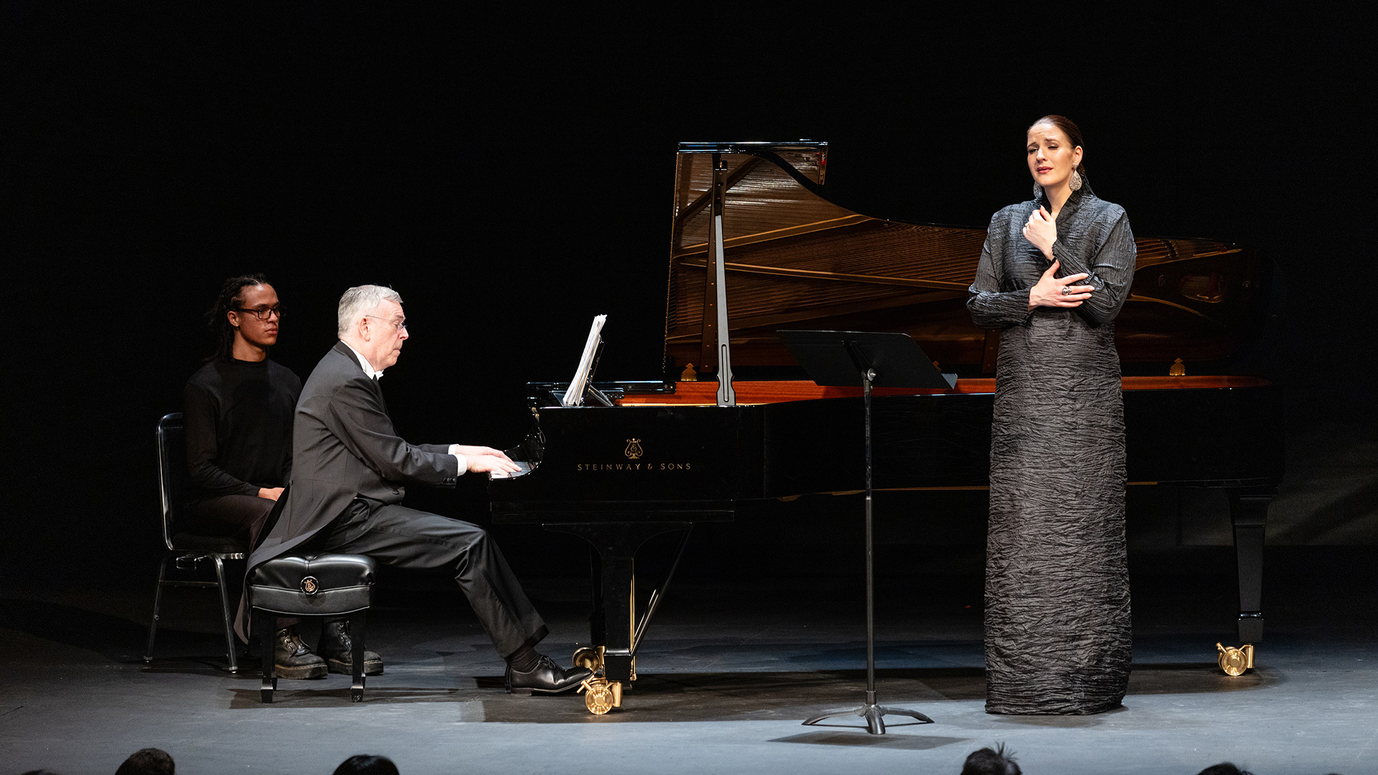 A woman in a charcoal floor length dress sings while a man in silhouette plays a grand piano