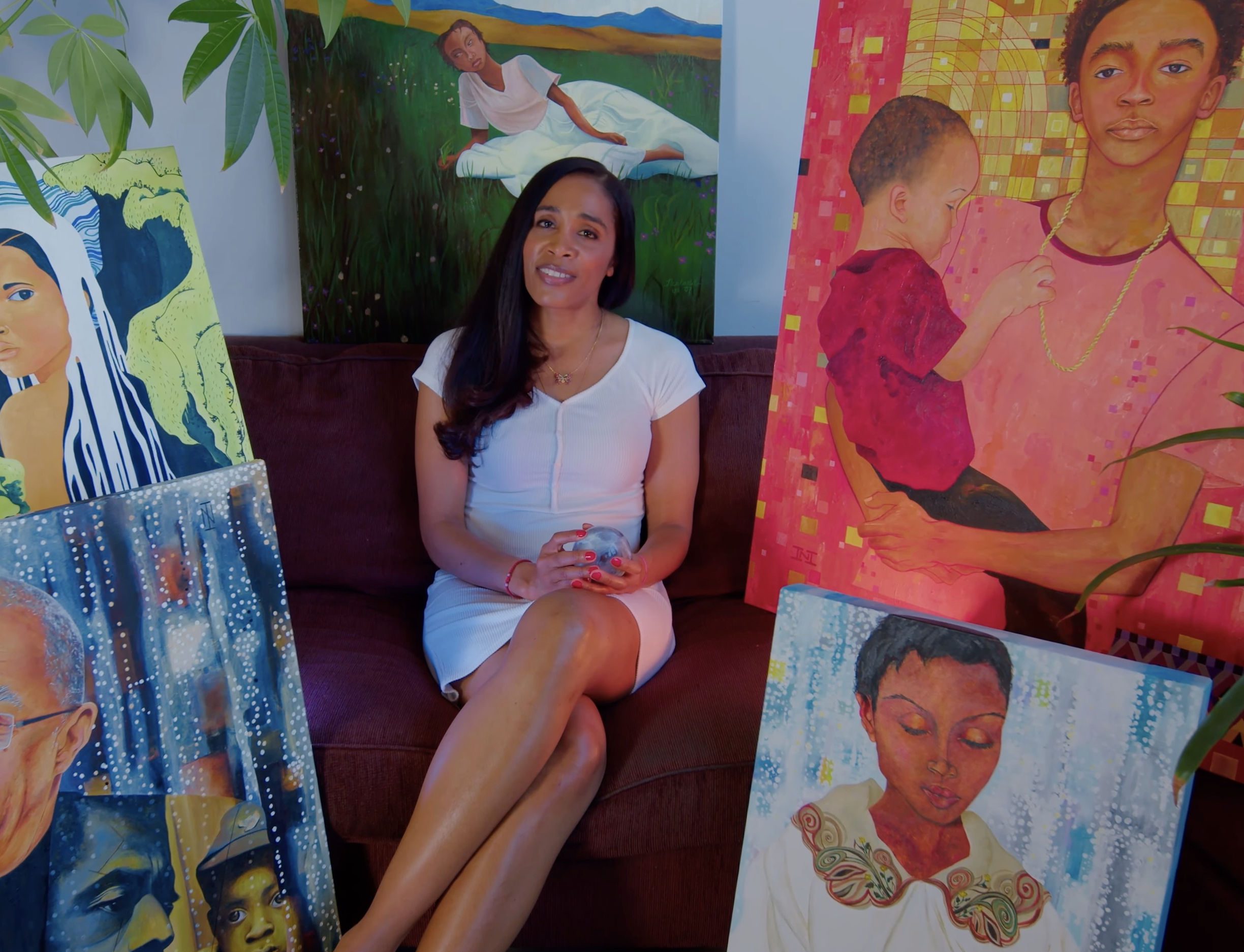 A woman sits on a couch, surrounded by painted portraits.
