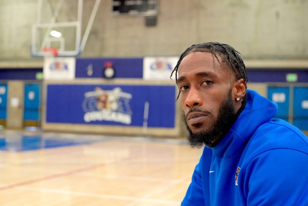 A Black man in a blue hoodie and beard looks at the camera with a basketball hoop and court in the background