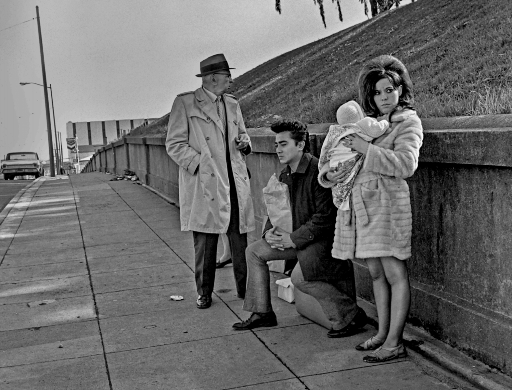 A young woman with backcombed hair stands in fur coat and flat shoes clutching a baby. Behind her a young man with a pompador crouches. Behind him is an older man wearing a raincoat and hat.