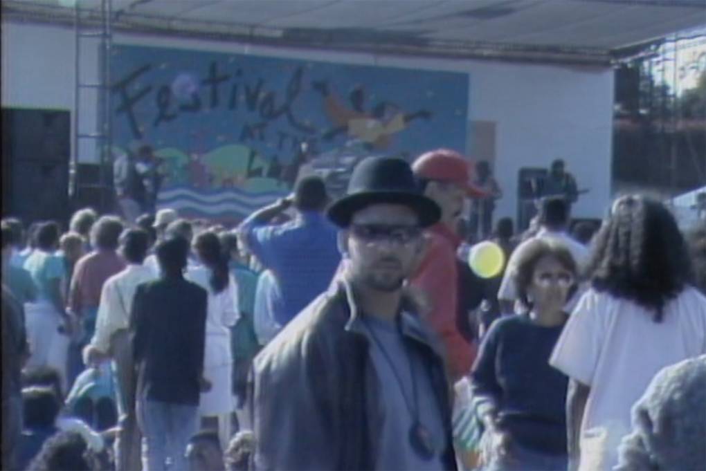 A man in a black derby hat and leather jacket looks at the camera, surrounded by people, with a stage in the background