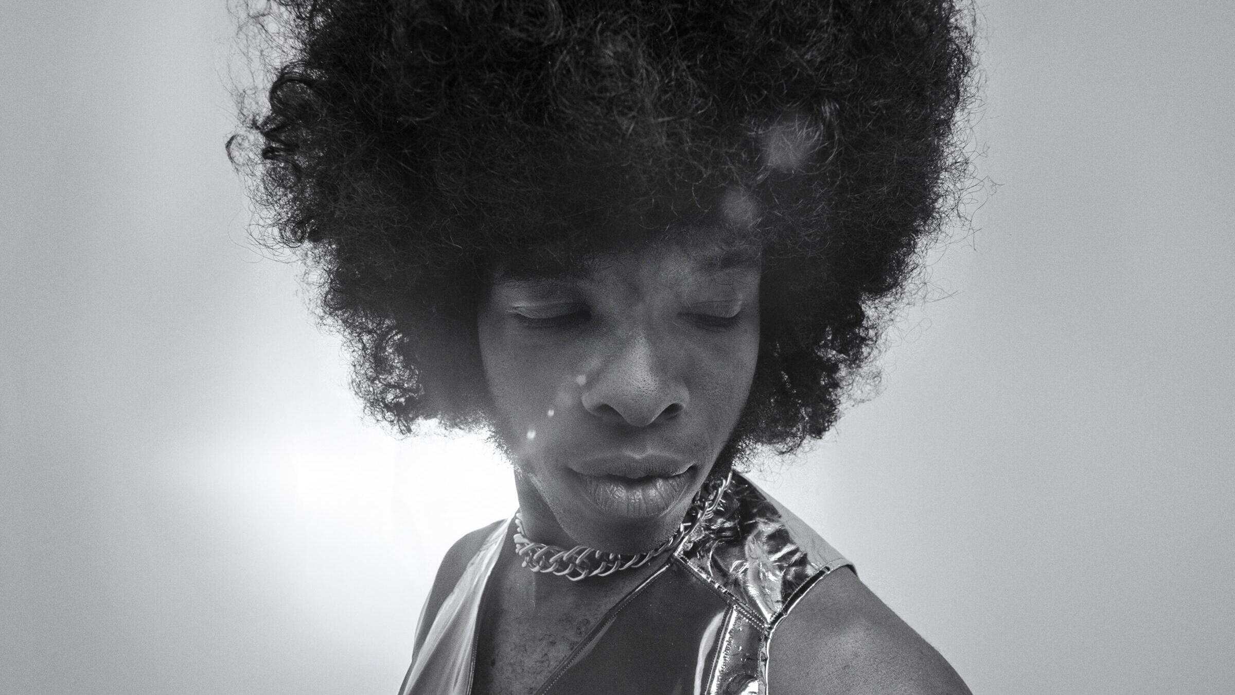 a Black man in a large afro and decorative necklace and shiny top, in a black and white photo
