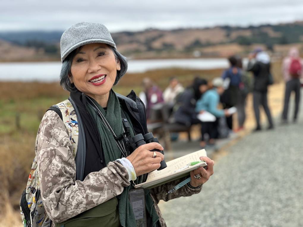 older Asian woman in hat smiles with notebook and binoculars in front of watery landscape