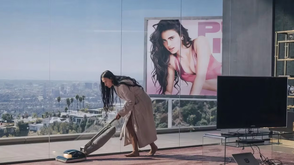 A womon vacuums a room with large glass walls overlooking the big city below, near a billboard featuring another younger woman
