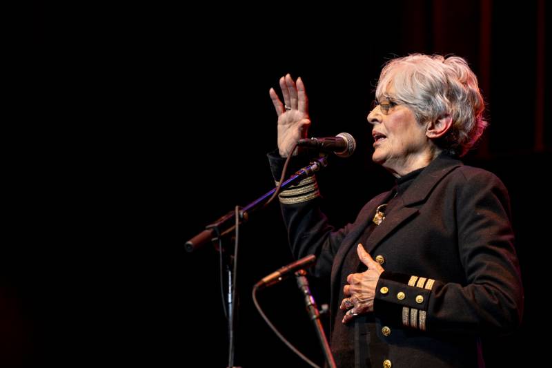 A woman in a fashionable black military-style jacket sings into a microphone, facing left, with her right arm in the air