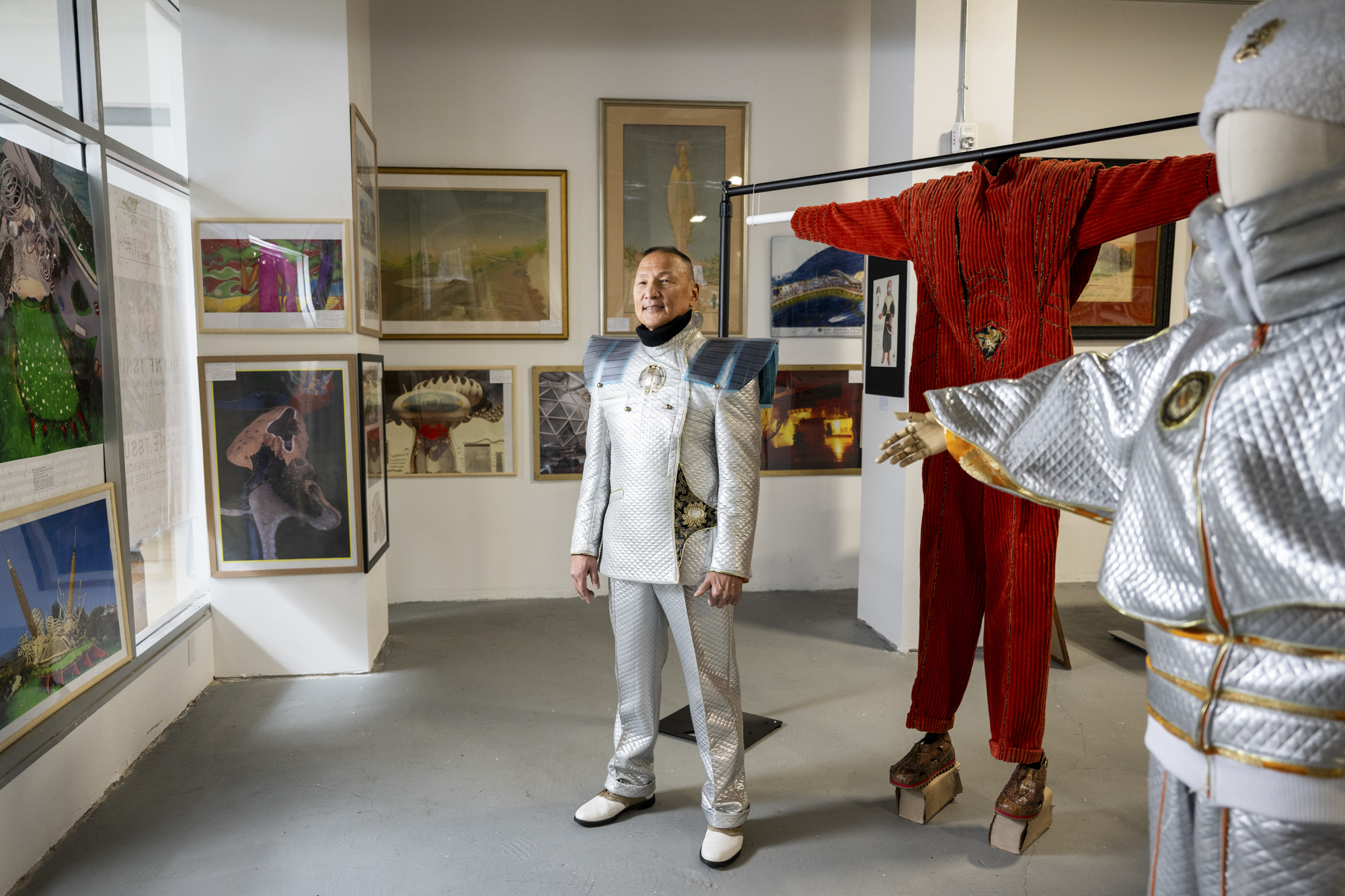 older man in silver outfit stands within gallery space filled with art and fashion