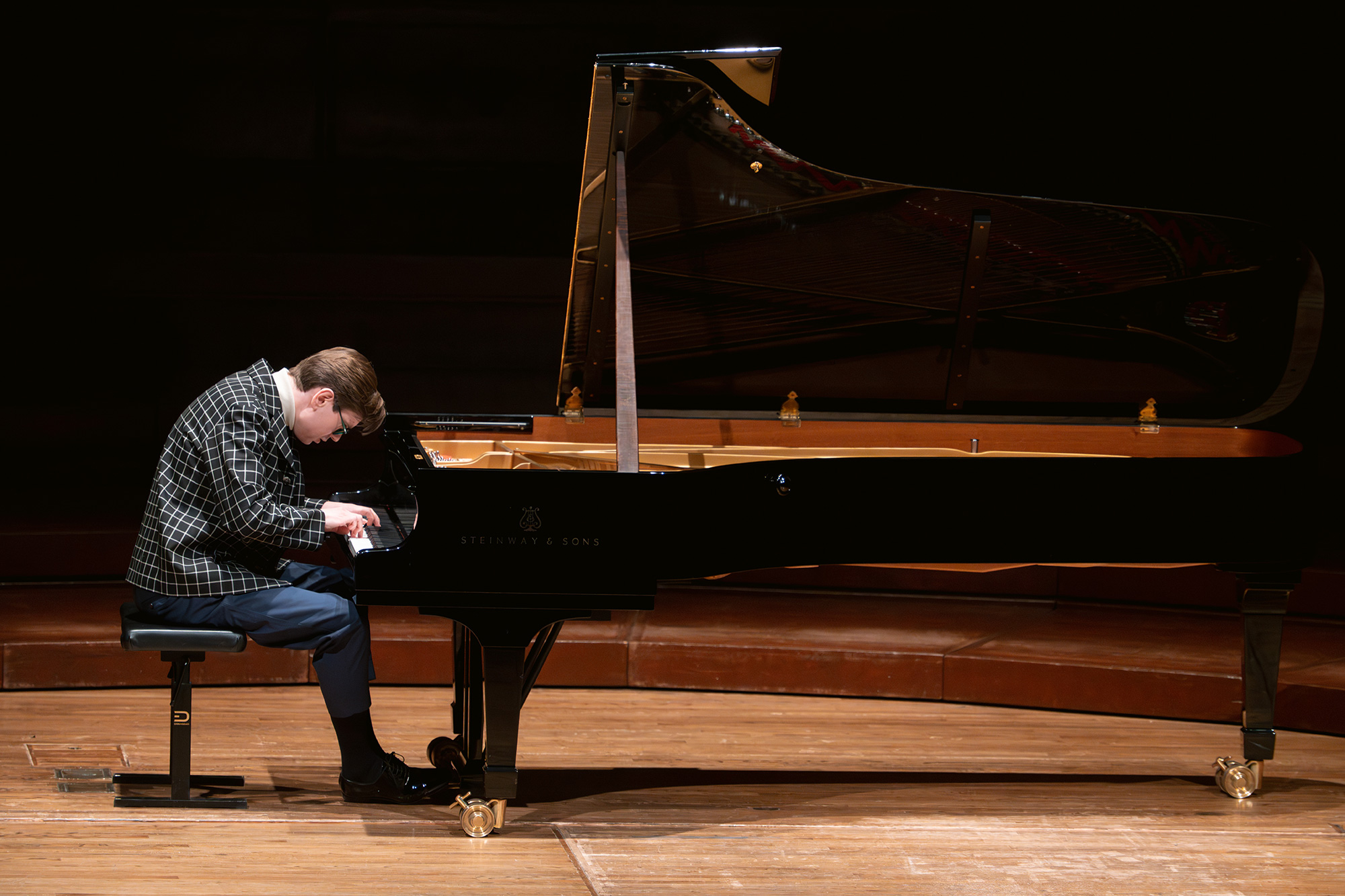 A man in blue slacks and patterned jacket leans over a grand piano keyboard on a sparsely lit stage