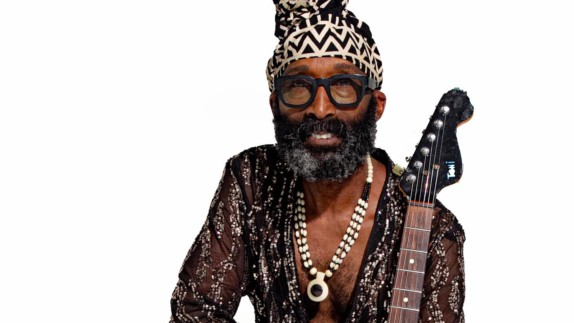 A man in glasses holding a guitar in a necklace and decorative shirt