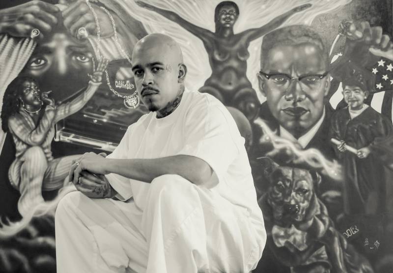 black-and-white image of man with shaved head in all white in front of detailed figurative artwork