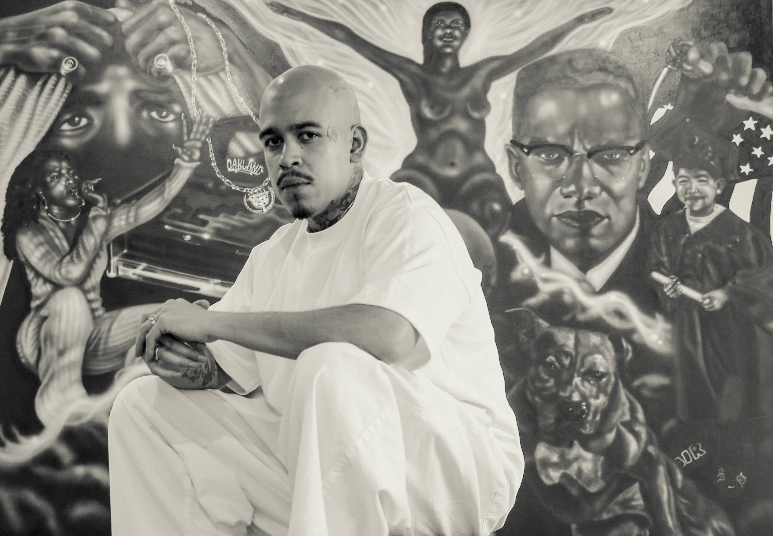 black-and-white image of man with shaved head in all white in front of detailed figurative artwork