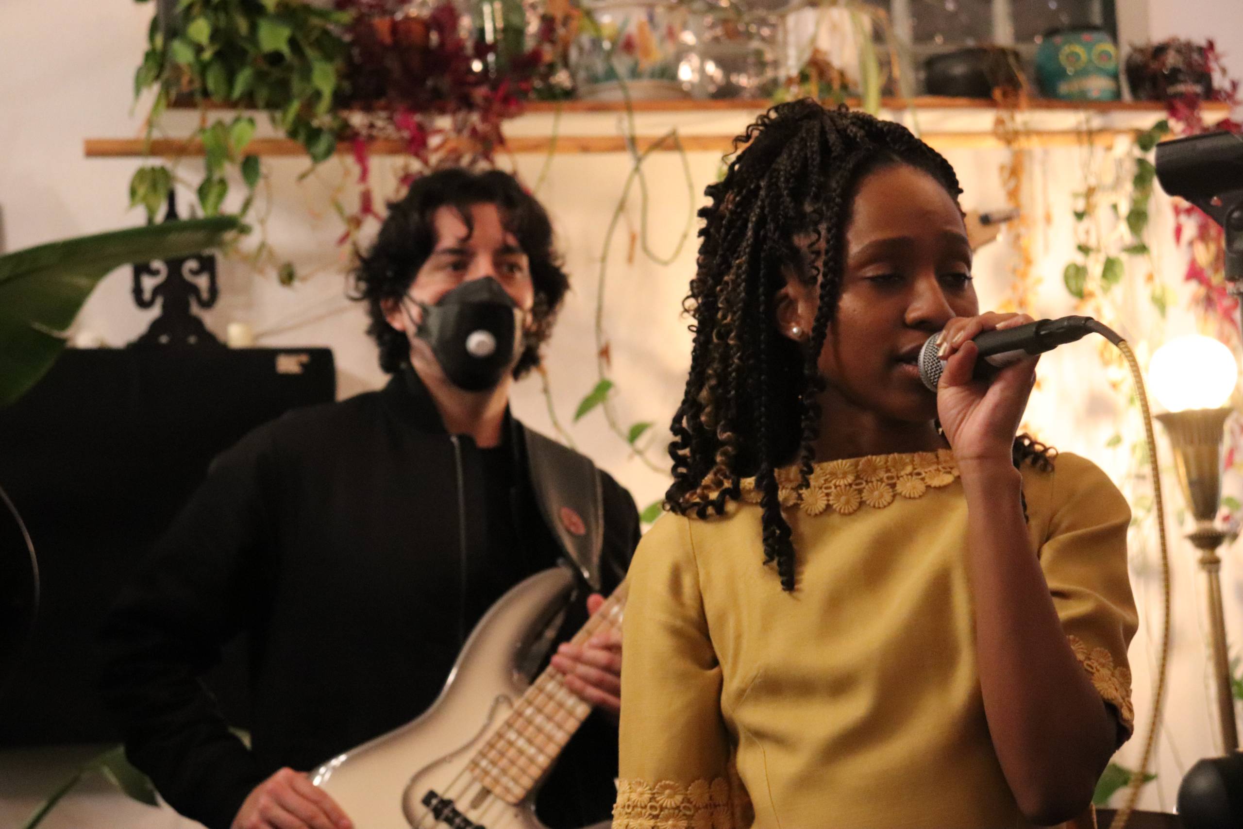 A singer sings into a microphone while a guitarist, who is wearing an N95 mask, plays behind her.