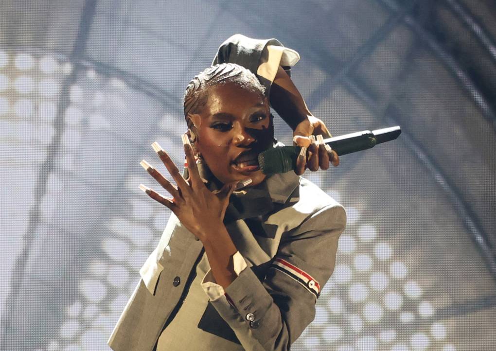 A young Black woman holds a microphone around the back of her head with lights in the background