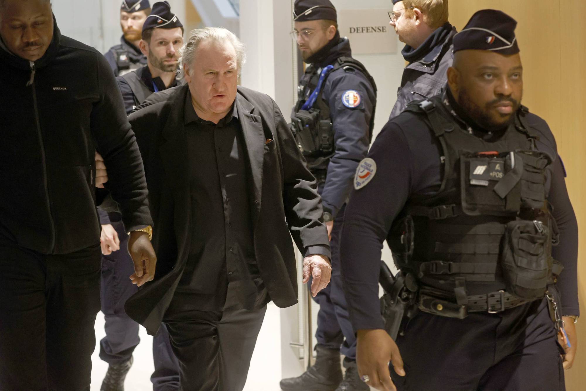 An older, rotund man with grey thinning hair walks through a corridor lined with French police officers. He is wearing a black suit.