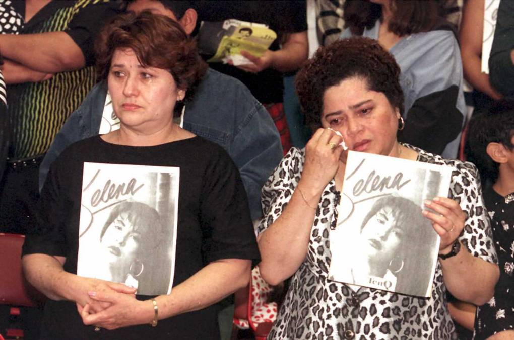 Two Latinas standing side-by-side holding up black and white photos of Selena. The woman on the right is wiping away a tear.
