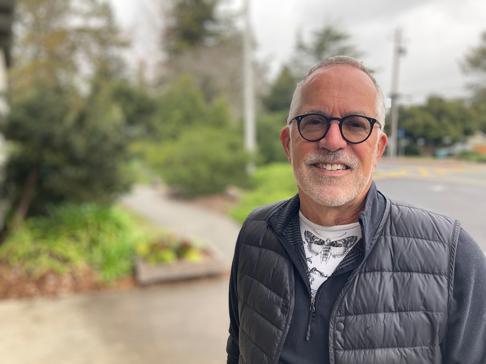 A man in glasses and a greyish puffed vest looks into the camera, smiling, against a backdrop of trees