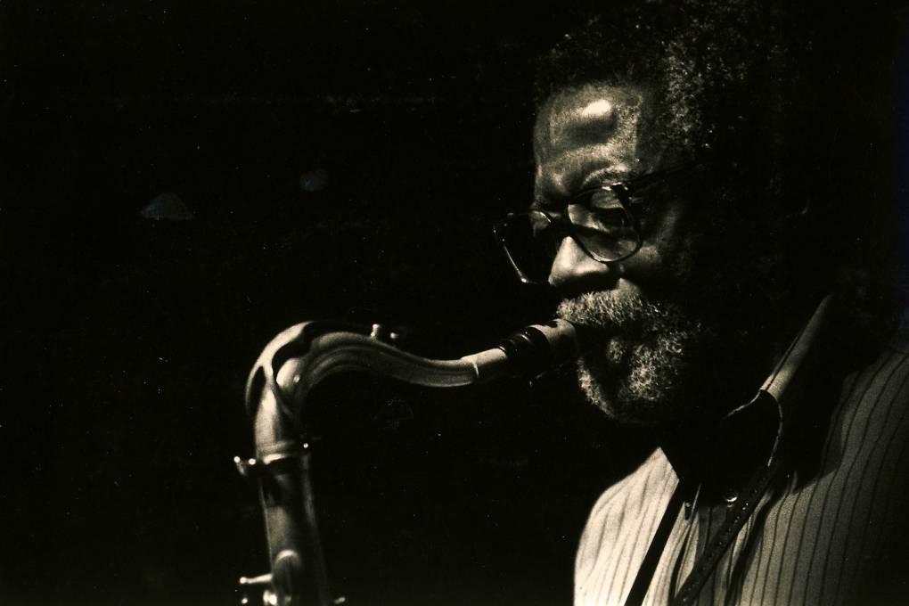 A Black man in glasses and beard plays a saxophone, facing left