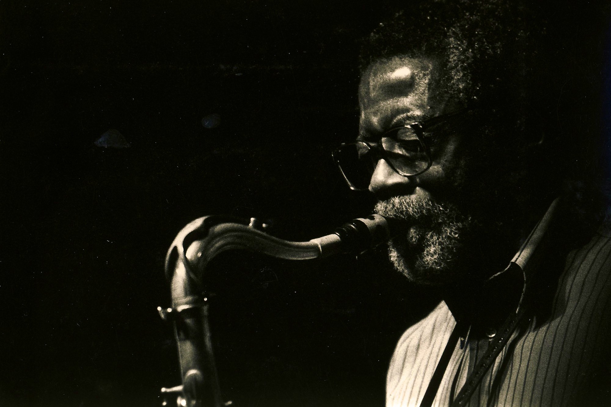 A Black man in glasses and beard plays a saxophone, facing left