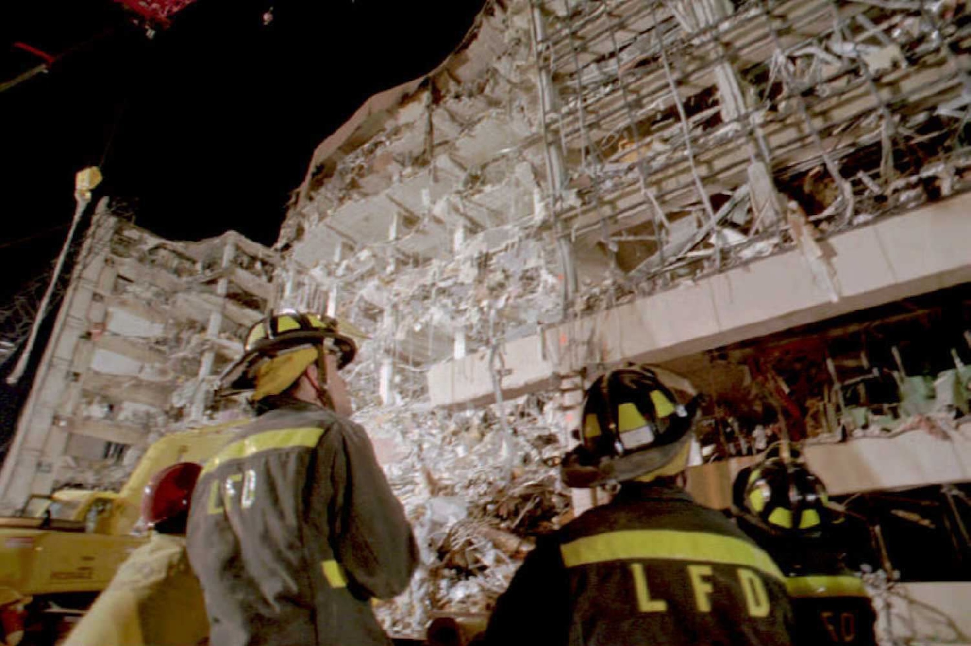 A half-destroyed building stands with its interior visible to the outside. Firefighters in uniforms marked LFD stand examining the wreckage from the ground.