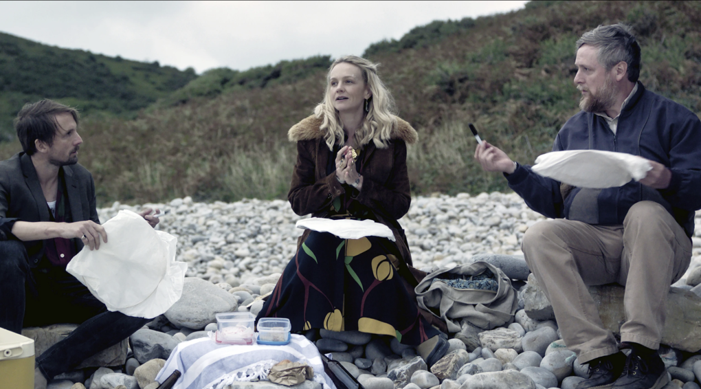 A pretty woman with long blonde hair sits on a pebble beach looking wistful. At her right is a man holding up a pen and papers. At her left is a frustrating looking man, also holding papers.