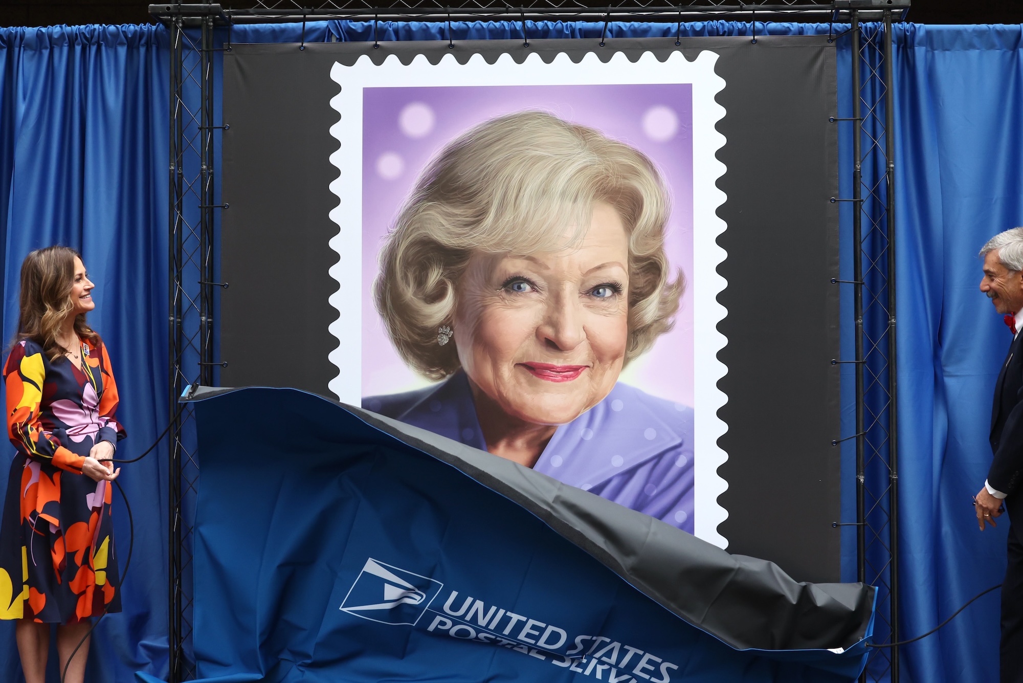 A large scale postage stamp featuring an illustration of Betty White is unveiled in front of blue curtains. A woman and man stand either side, admiringly.