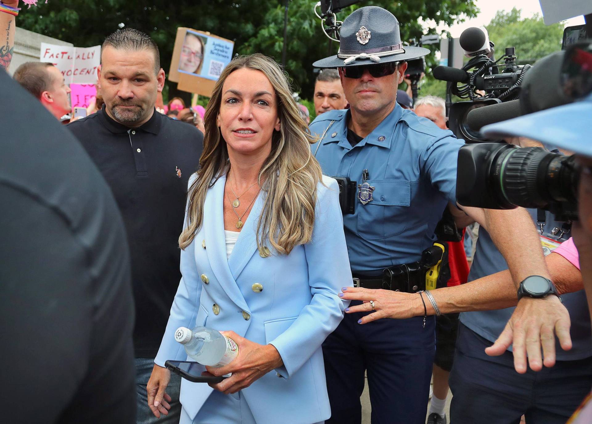 A slender woman with long blonde hair, wearing a powder blue suit, walks through crowds holding signs, flanked by law enforcement officers.