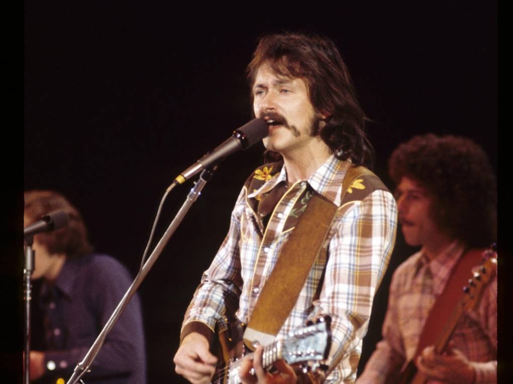 A man with an excellent 1970s hairdo and mustache plays guitar with a brown leather strap and briwn flannel shirt while singing into a microphone