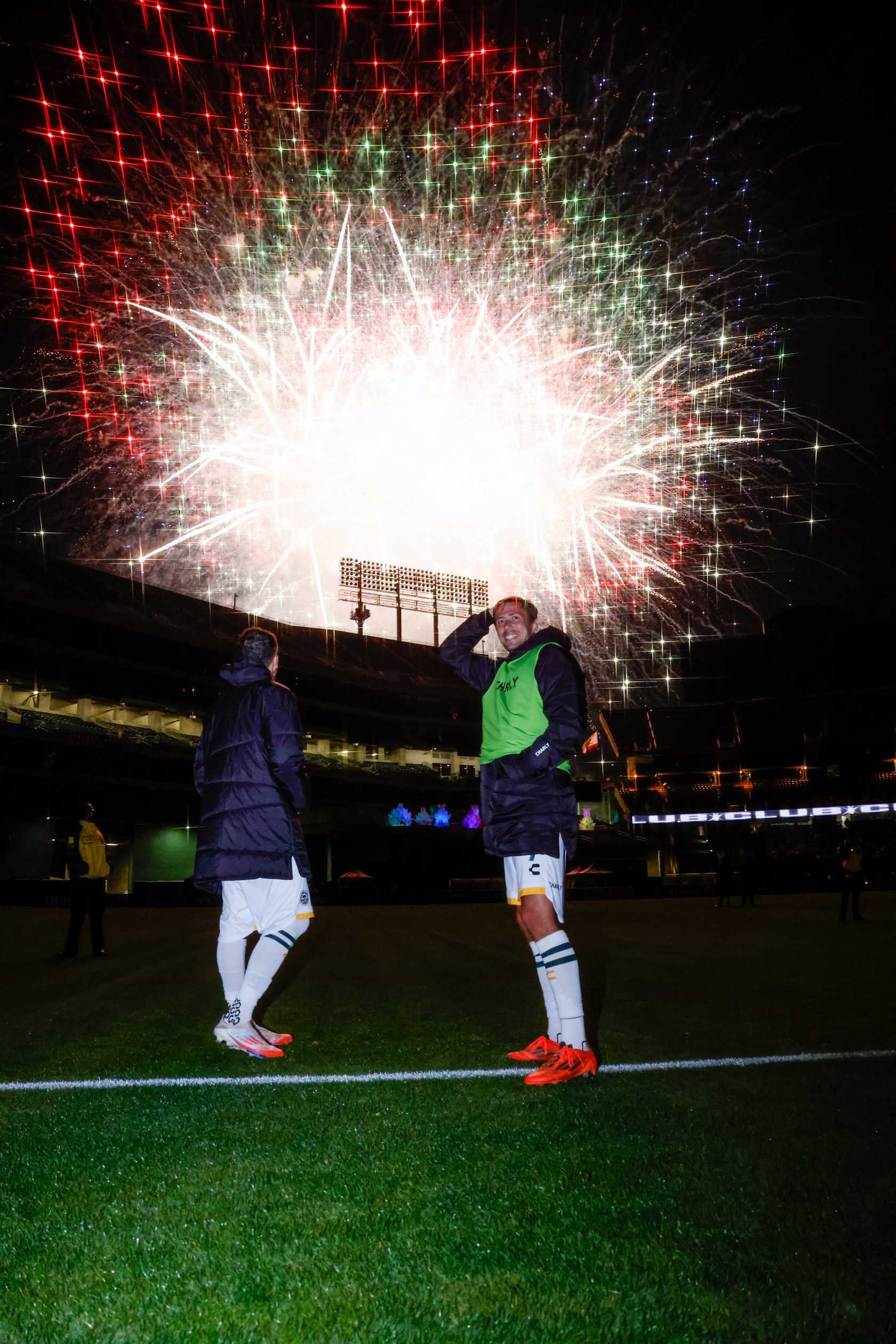 After a soccer game two players stand on a dark field as fireworks light up the sky above.