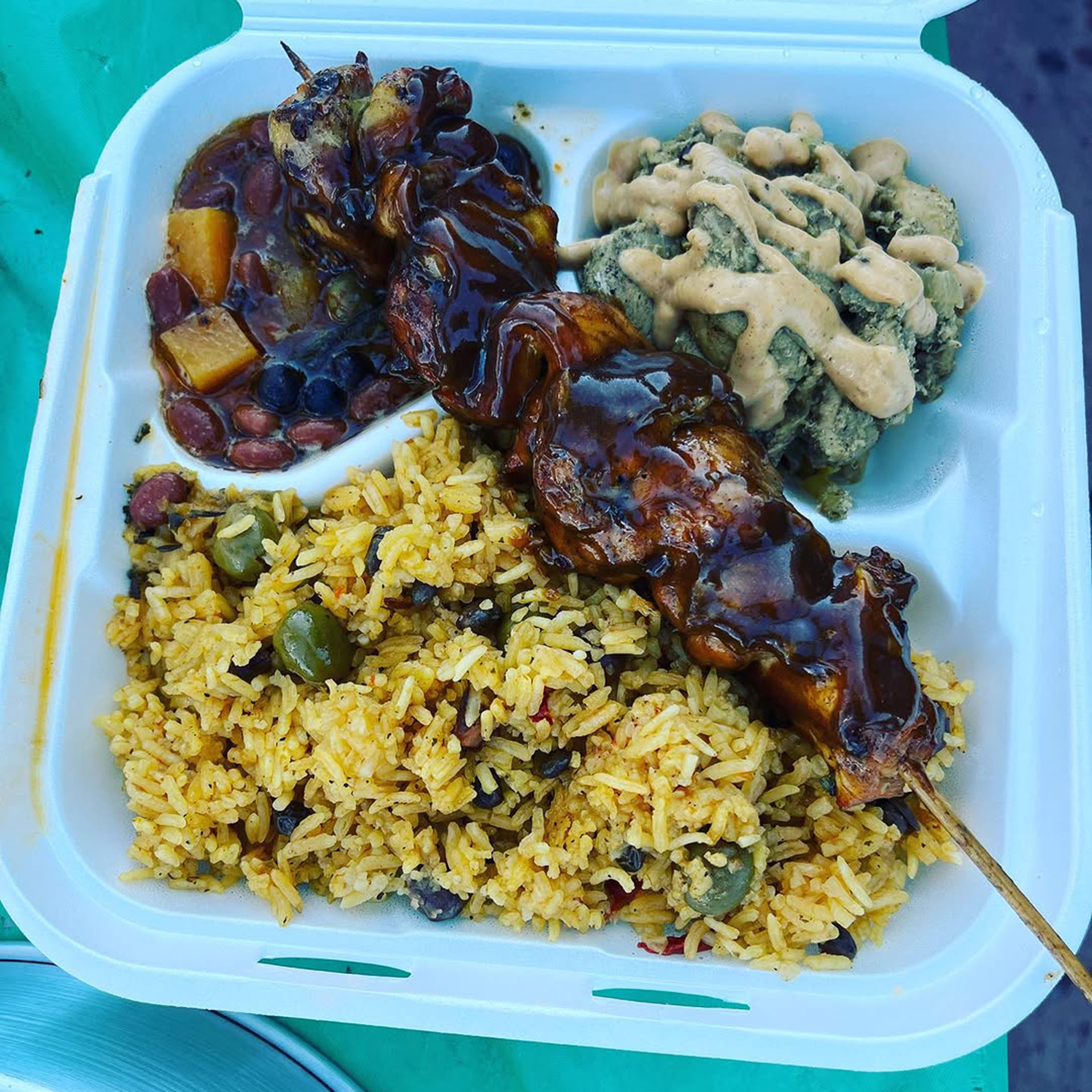 A takeout container with rice, beans, and skewered meat.