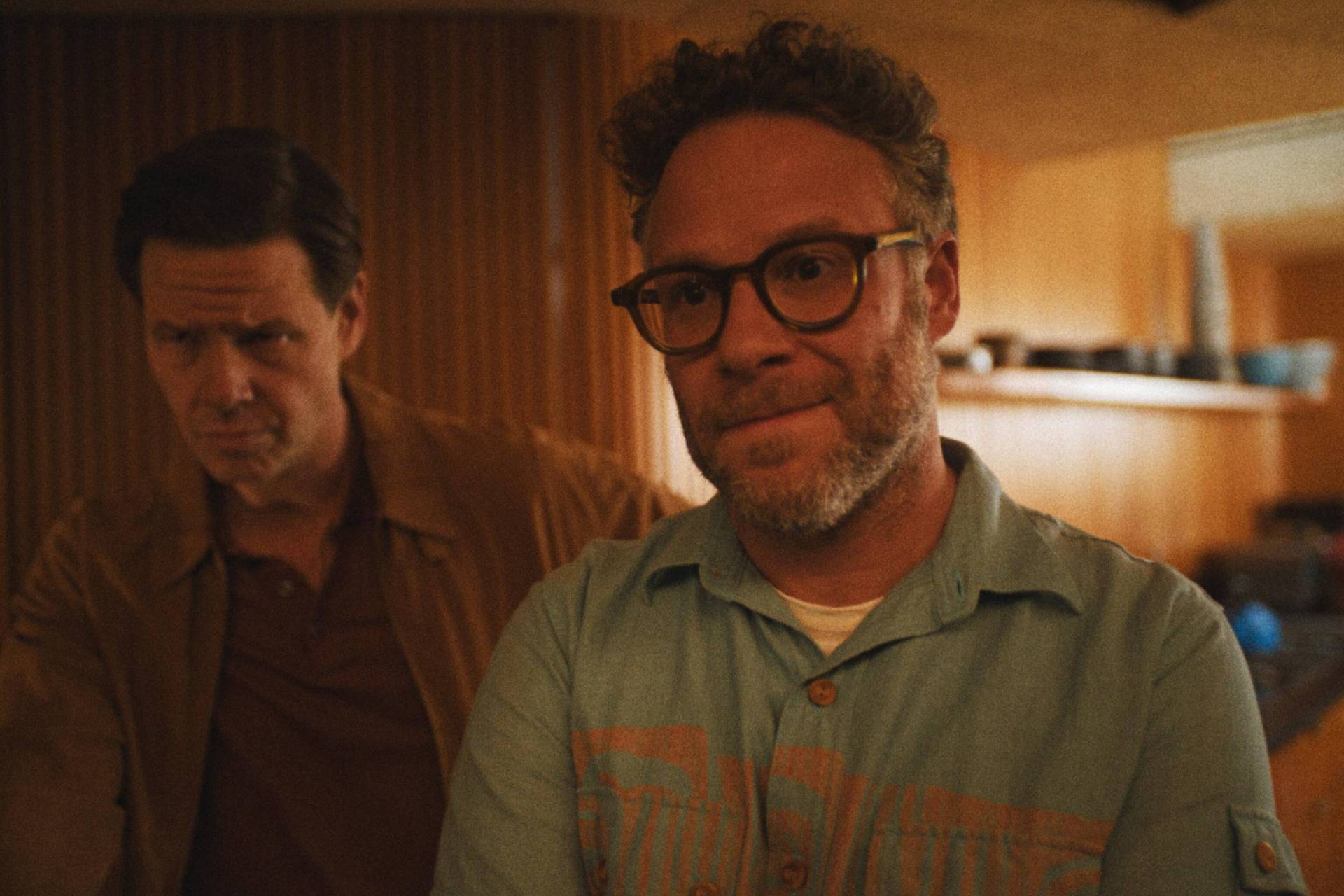 A middle-aged man wearing a casual blue shirt and round glasses stands in a wood-paneled room watching something proudly. A frowning man stands near him, looking more cynical.