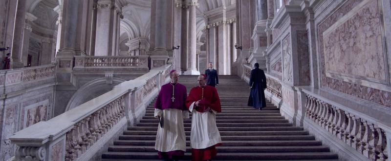 Two cardinals in white robes and red capes walk down grand stone stairs in a huge stone building.
