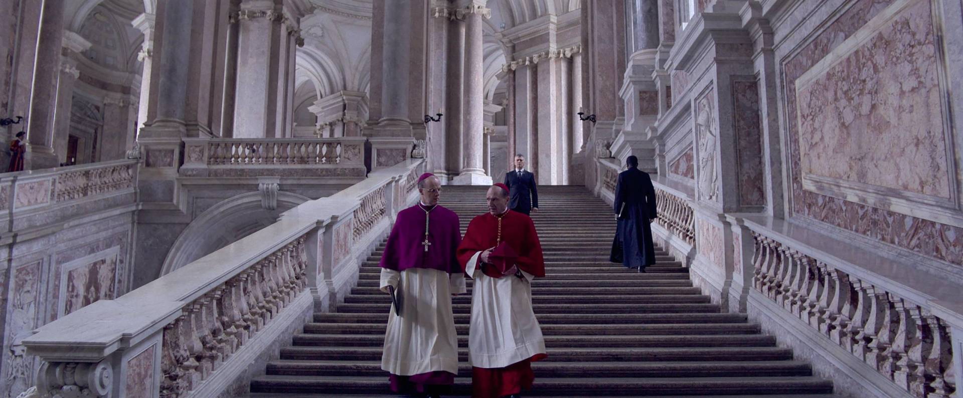 Two cardinals in white robes and red capes walk down grand stone stairs in a huge stone building.