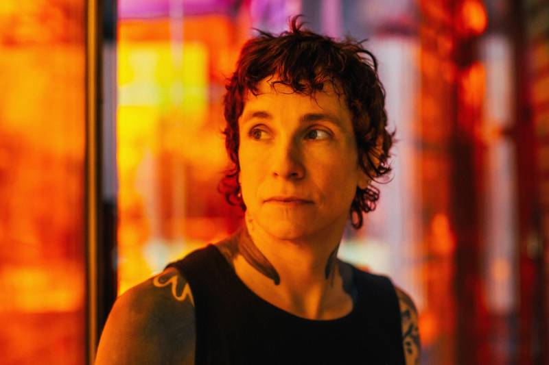 a portrait of a woman with short hair and a sleevless tank top with tattoos, looking pensive in front of a pink and orange background