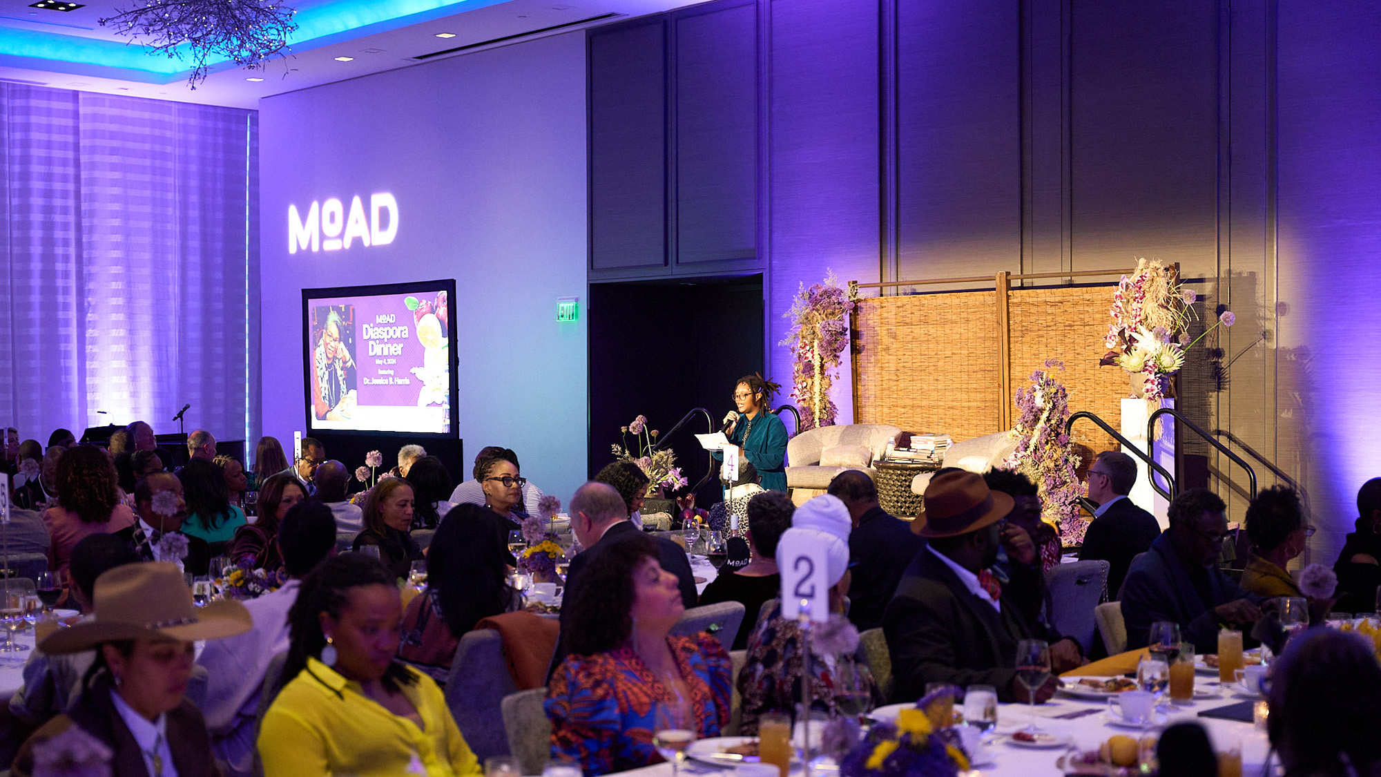 Guests seated at long tables for a fancy dinner banquet. The neon sign on the wall reads, 'MoAD'.