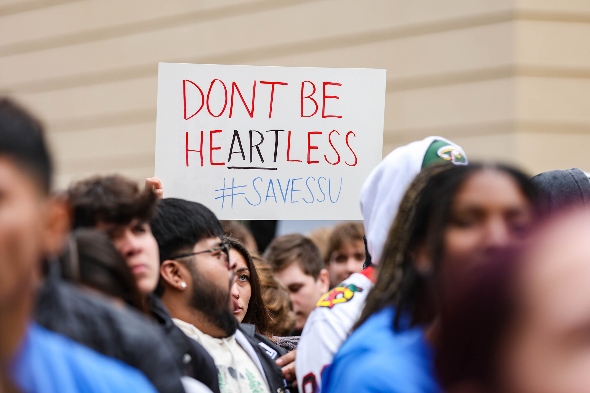 crowd with handmade sign that reads 'don't be heartless #savessu' with 'art' underlined
