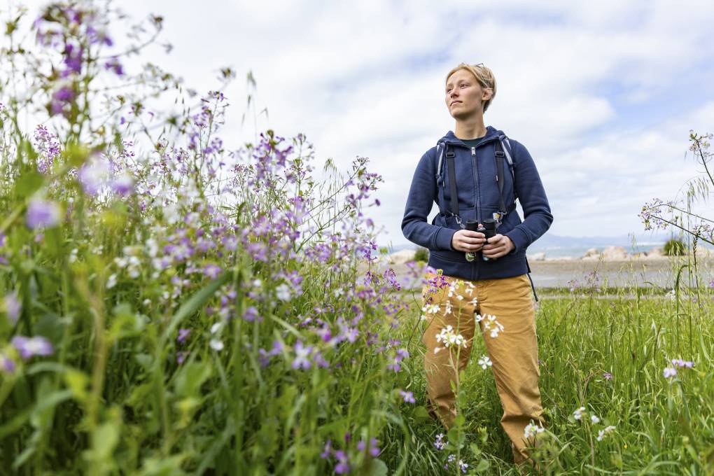 young person with short blonde hair stands in grassy field with binoculars
