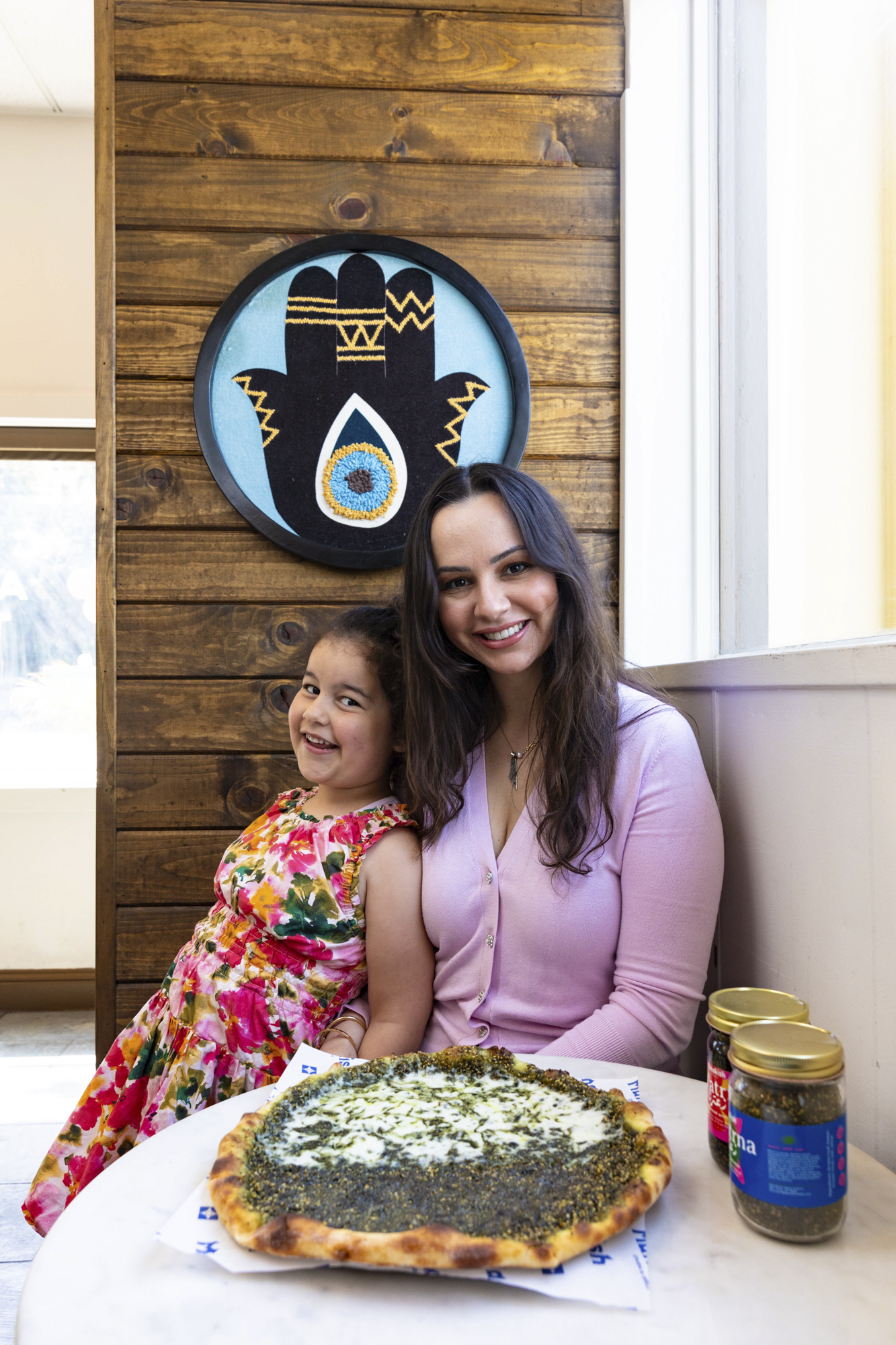 A mother and child pose for a portrait, seated inside a restaurant with a cheese-topped flatbread in front of them.