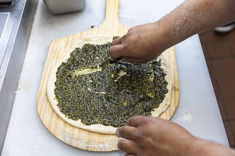 Bright green za'atar spice blend being spread on a round of dough.