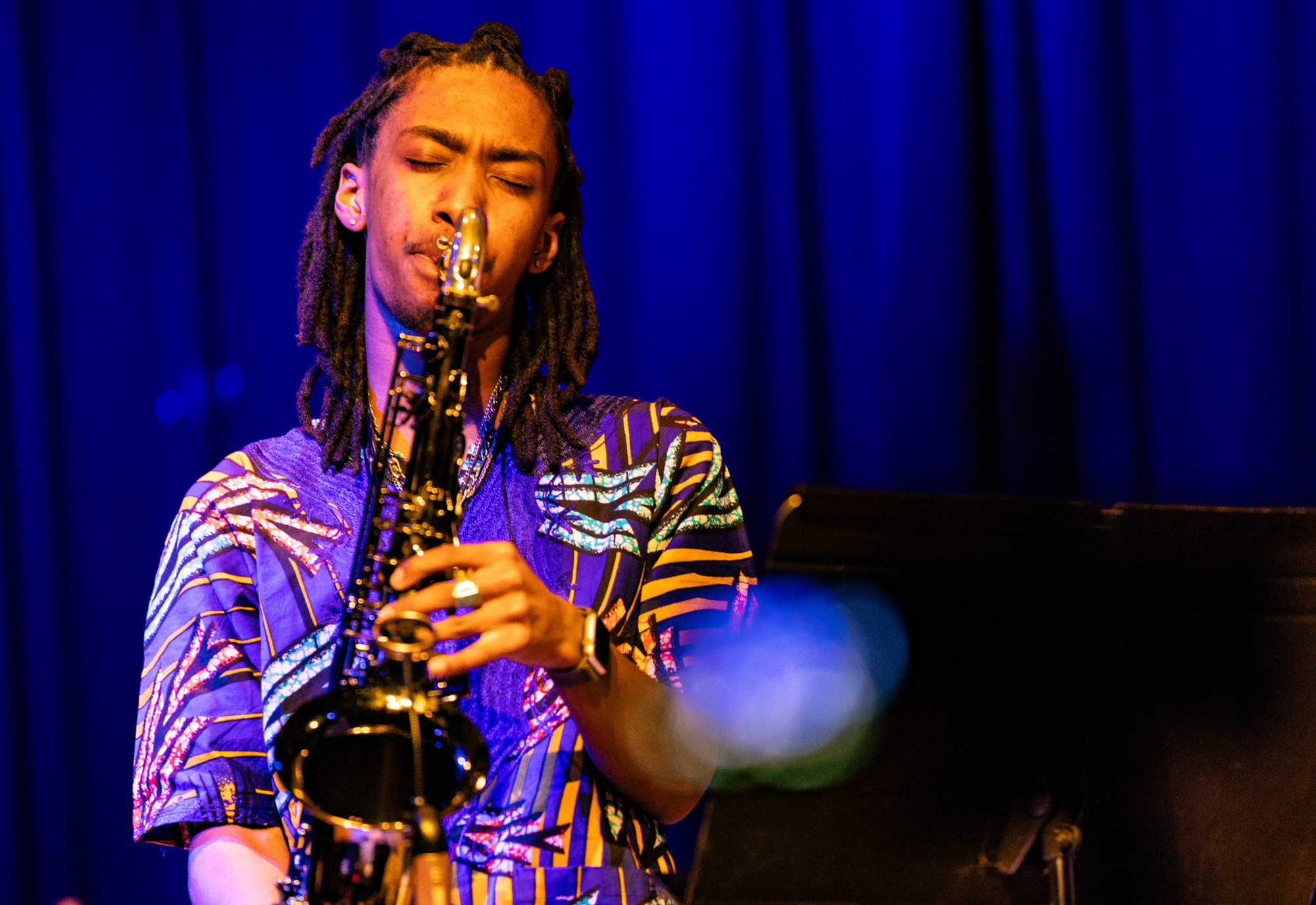 A young Black man in a patterned blue shirt plays the tenor saxophone with his eyes closed against a blue curtain background