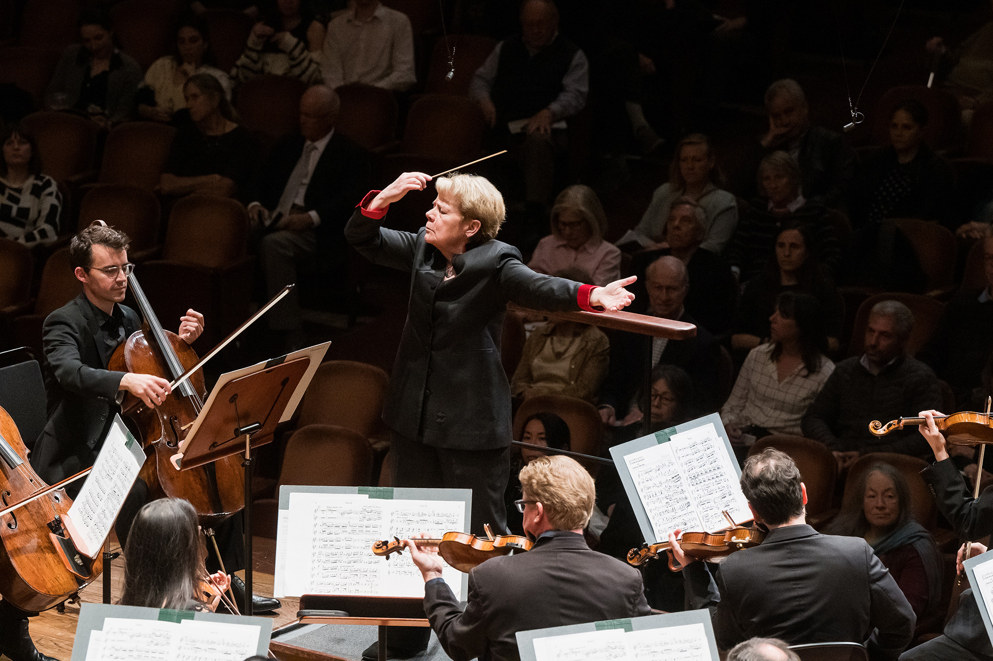 A woman in a dark suit and short hair moves her arms while conducting an orchestra, with the audience seen in the background