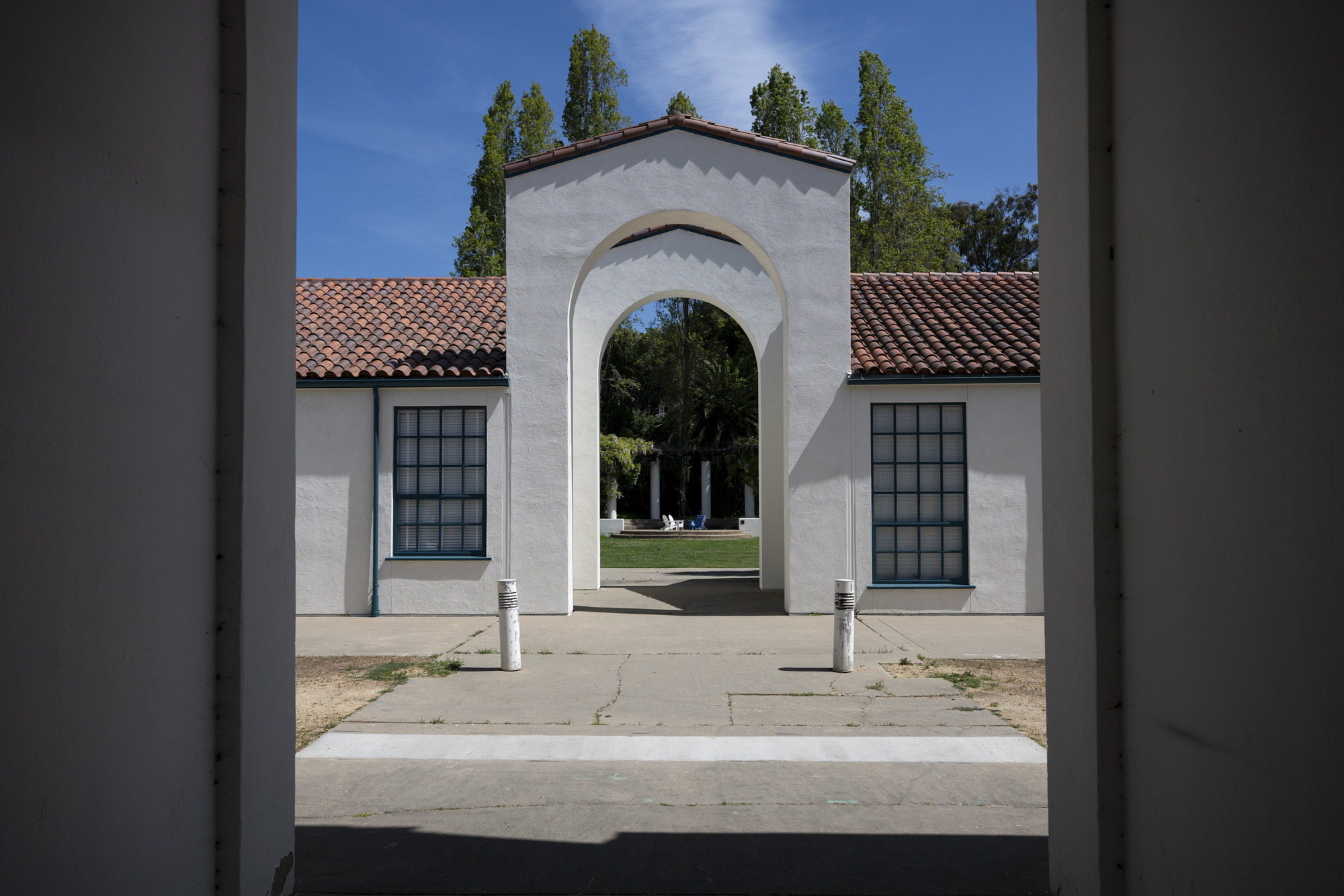 view through archway to mission-style buildings with tile roofs, empty chair on lawn