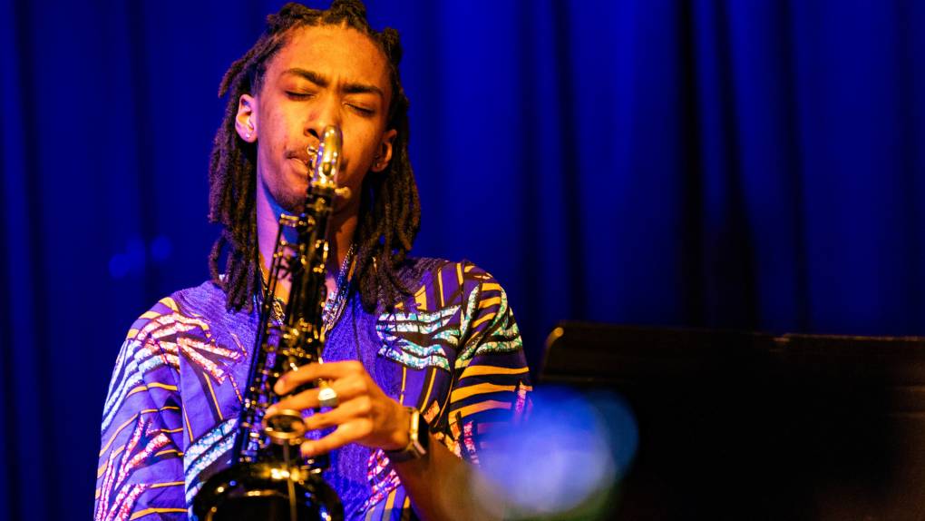 A young Black man in a patterned blue shirt plays the tenor saxophone with his eyes closed against a blue curtain background