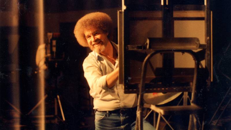 white man with afro peeks from behind easel while painting