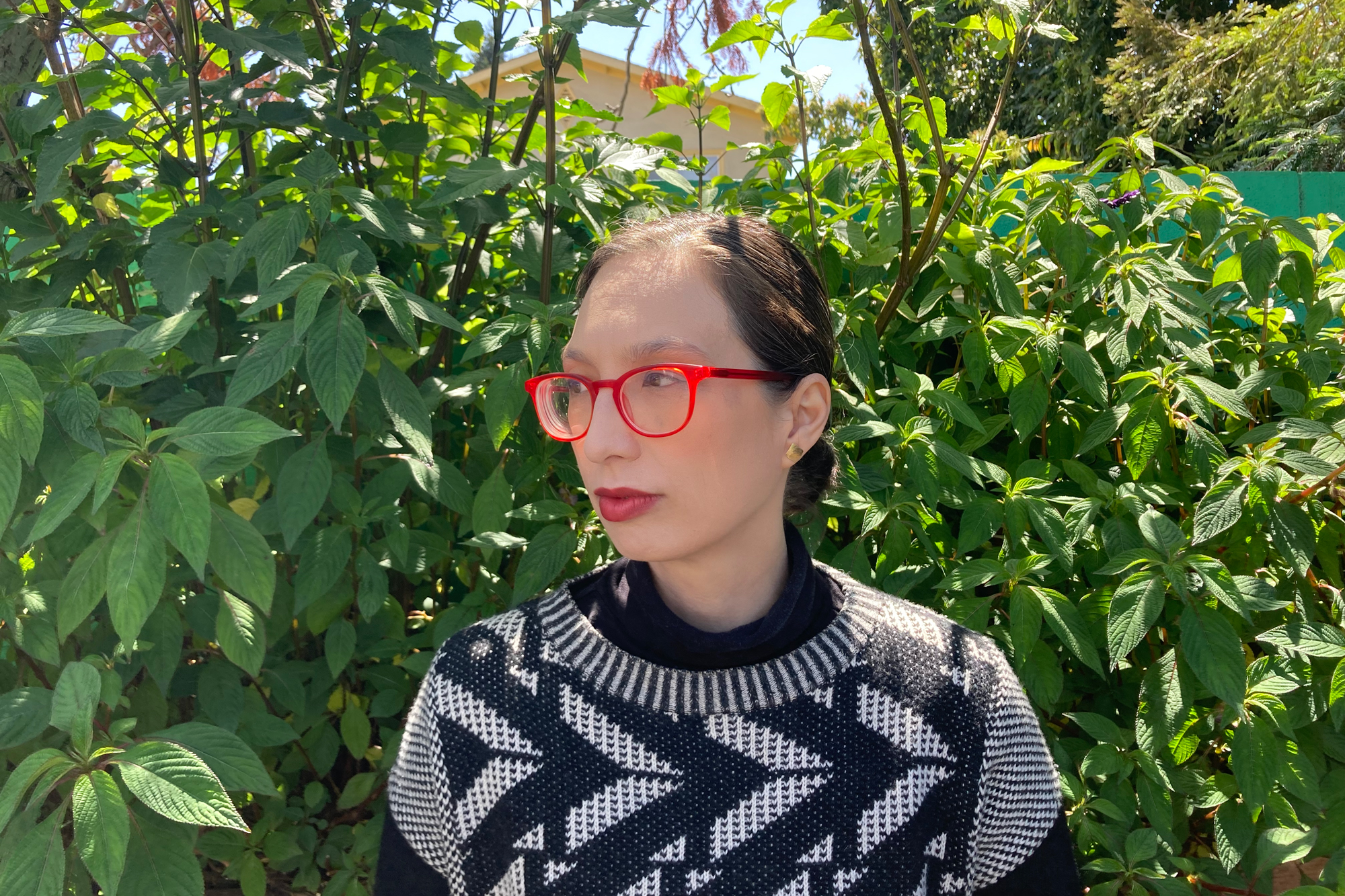asian woman with red framed glasses in sweater stands in front of bush