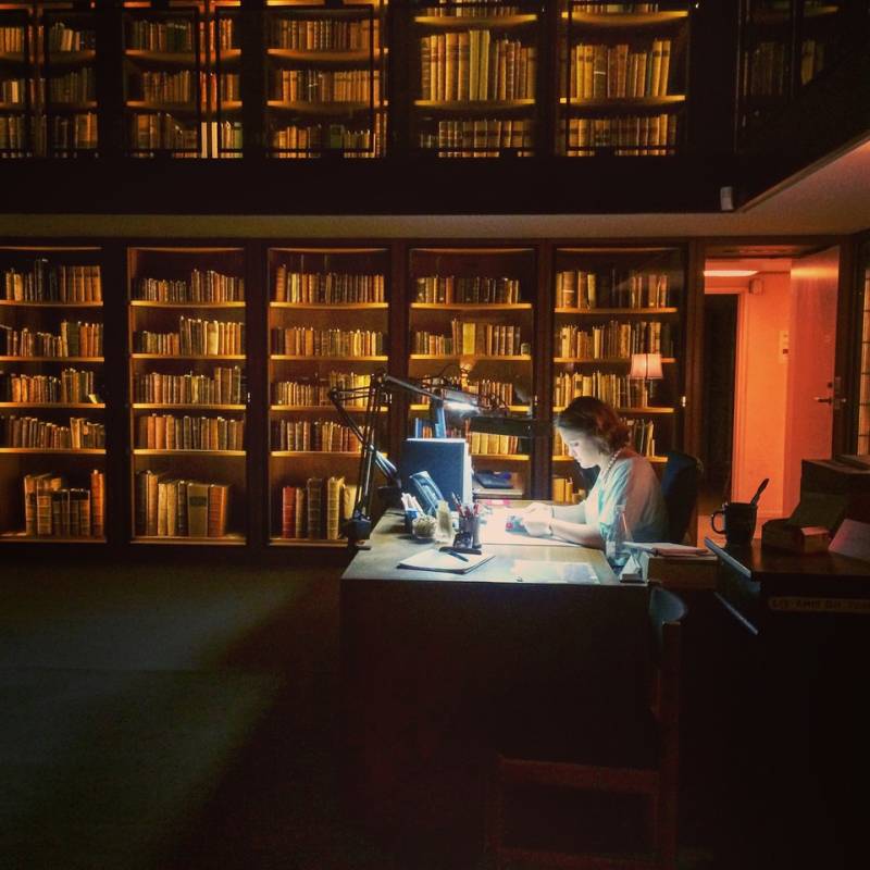 A young white woman sits at a desk, illuminated by a lamp, in a dark room surrounded by bookcases full of books