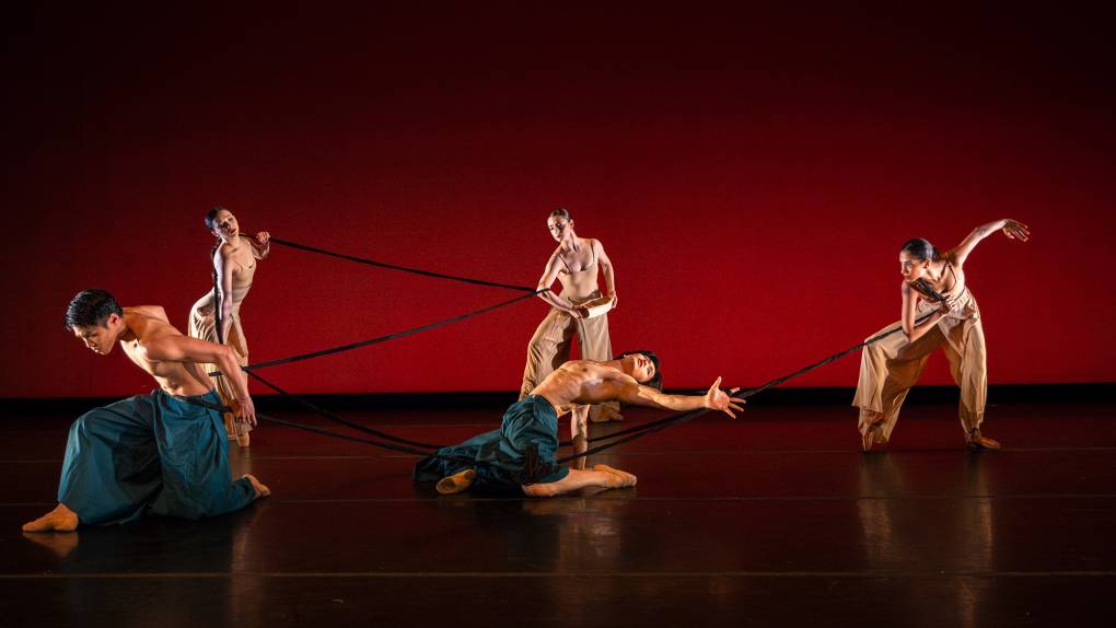 Four dancers, all Asian, perform against a deep red backdrop with a long braided plait.