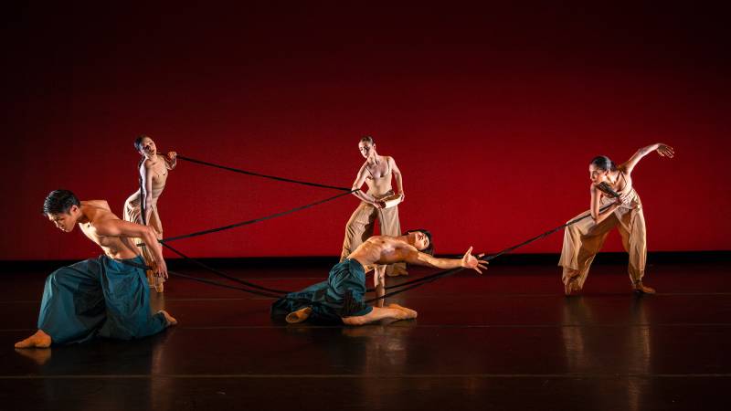 Four dancers, all Asian, perform against a deep red backdrop with a long braided plait.