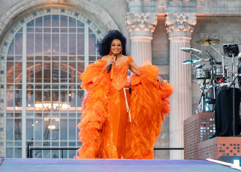 Diana Ross smiles and holds a mic as her tulle orange gown billows in the wind during an outdoor concert.